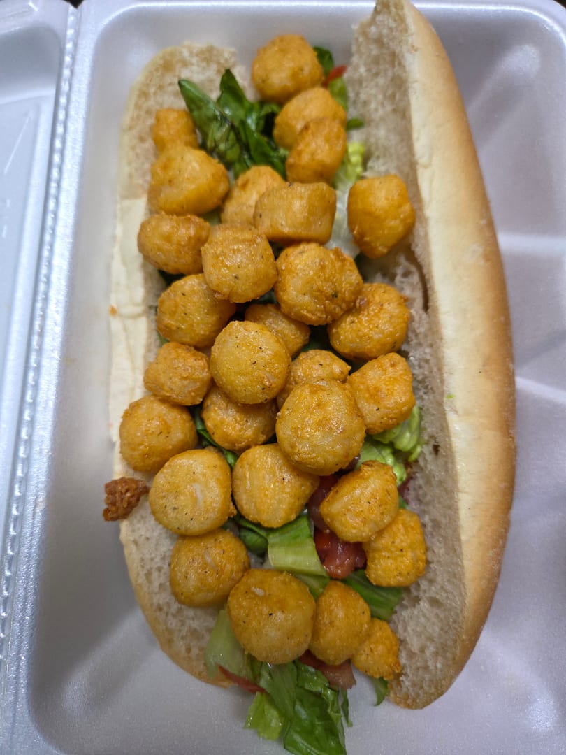 A close-up of a fried shrimp po'boy sandwich with lettuce and tomato in a white takeout container.