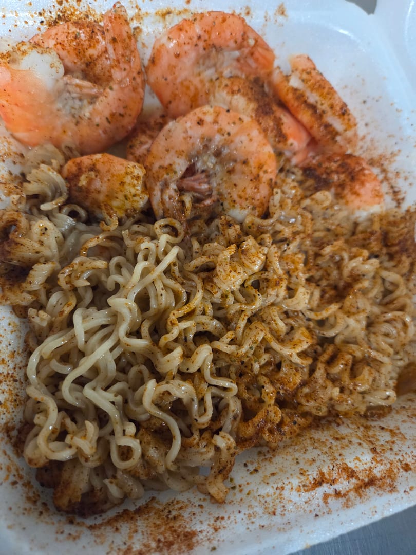 Spicy shrimp and ramen noodles, generously seasoned with red pepper flakes, in a white takeout container.