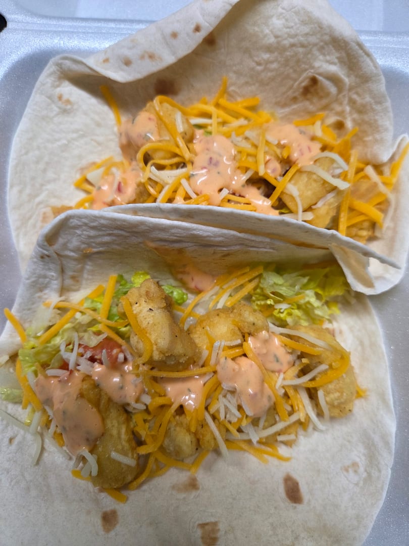 Two fish tacos with fried fish, shredded cheese, lettuce, and pink sauce in a white container.